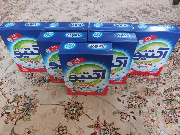 پودر لباسشویی اکتیو: بسته‌های آبی و سفید با نشان تجاری Active روی یک فرش طرح‌دار گل‌دار قرار گرفته‌اند.