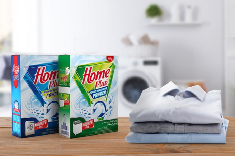 پودر لباسشویی ماشینی بدون فسفات ۵۰۰ گرمی هوم پلاس در کنار لباس و ماشین لباسشویی در یک خانه