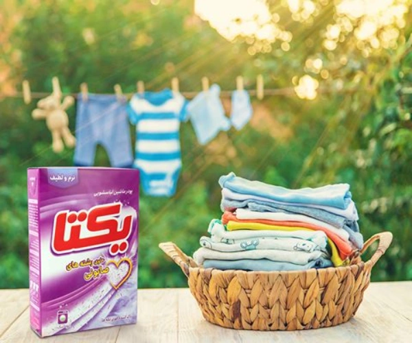 پودر لباسشویی یکتا ۵۰۰ گرمی با رشتههای صابون طبیعی، بستهبندی بنفش کنار سبد لباسهای رنگی کودک در فضای سبز با لباسهای آویزان