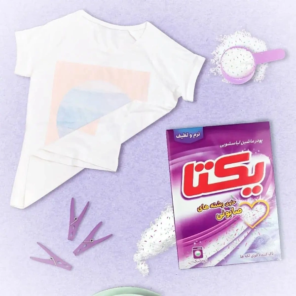 پودر لباسشویی یکتا ۵۰۰ گرمی با رشته های صابون، بسته بندی بنفش، کنار لباس سفید، گیره لباس و قاشق اندازه گیری روی زمینه بنفش